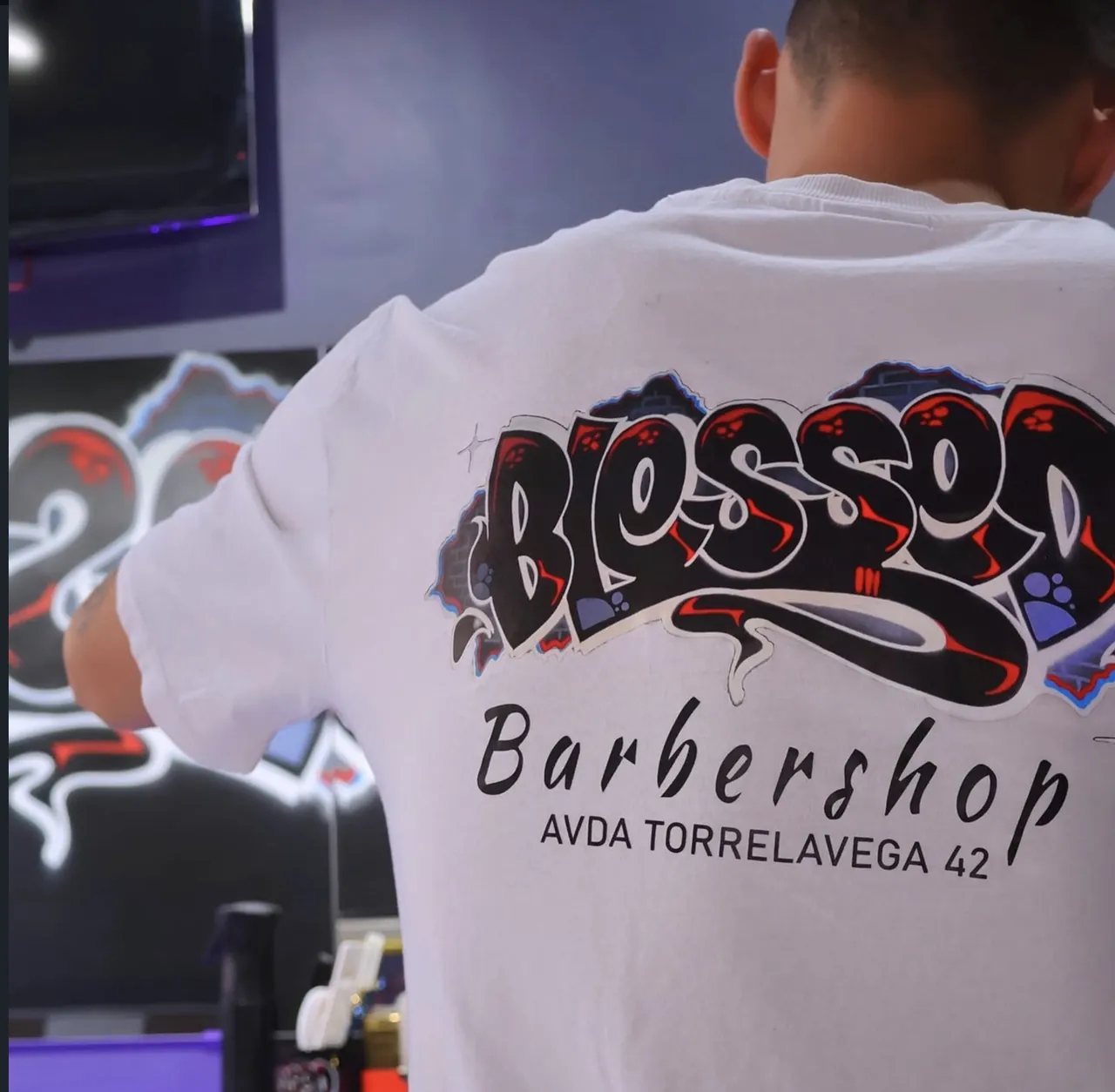 Espalda de una camiseta blanca con el logotipo de Blessed Barbershop y la dirección Avda Torrelavega 42