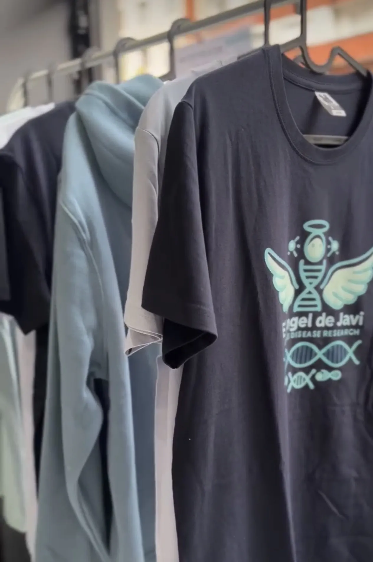 Camiseta azul marino con el logotipo de El Ángel de Javi, colgada junto a otras prendas en una barra